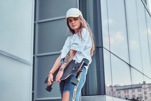 Premium Photo | Young Hipster Girl In Cap Dressed In White Shirt And Ripped  Jeans Holds Skateboard While Posing Near Skyscraper.